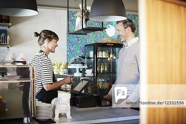 Smiling barista talking to customer while using cash register at checkout counter in cafe