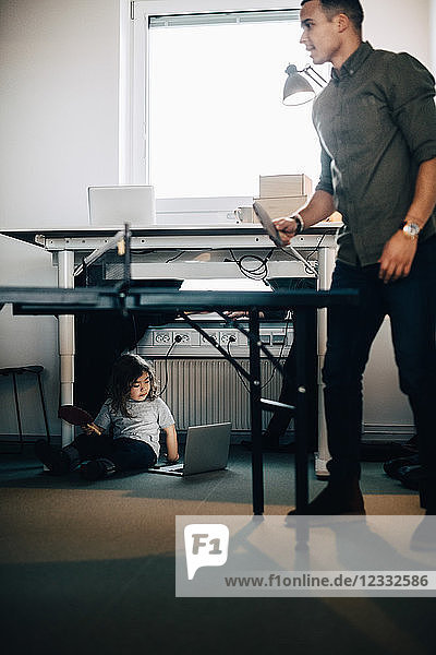 Full length of businessman playing table tennis while boy using laptop at creative office