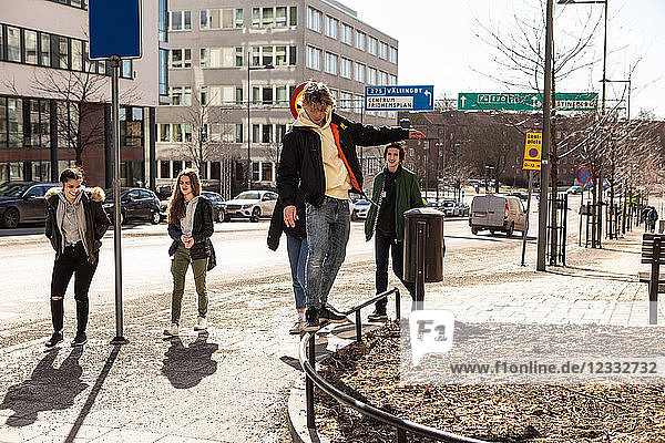 Teenage boy balancing on railing while friends walking on sidewalk in city during sunny day