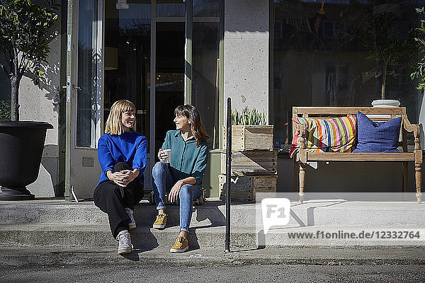Full length of smiling owners talking while sitting on steps outside store