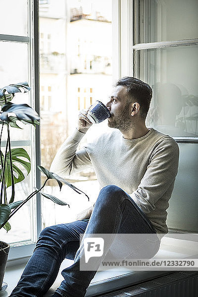 Thoughtful young man drinking coffee while sitting on window sill