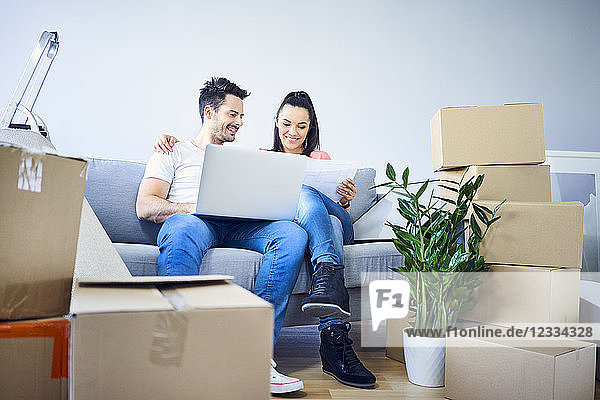 Happy couple sitting on couch surrounded by cardboard boxes using laptop