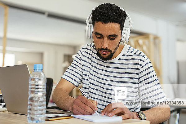 Young man wearing headphones taking notes at desk in office