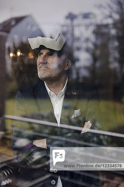 Senior businessman standing at window with arms crossed  wearing VR goggles