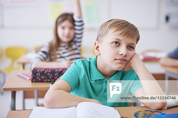 Bored schoolboy in class with girl raising her hand in background