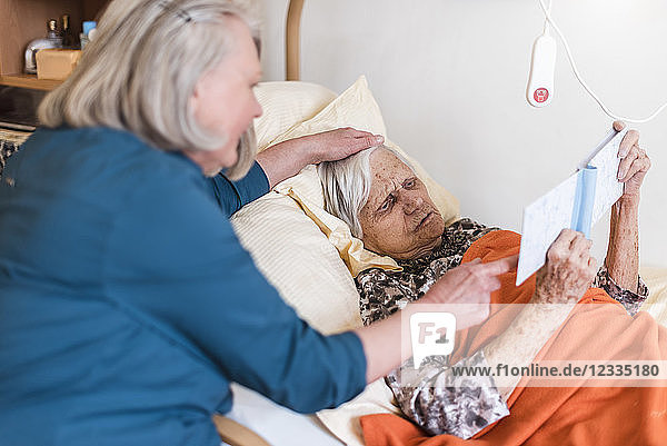 Woman taking care of old woman lying in bed reading book
