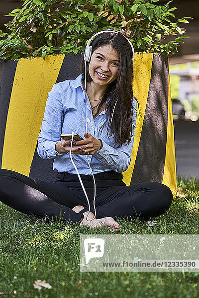 Portrait of smiling woman sitting barefoot on a meadow listening music with headphones and cell phone