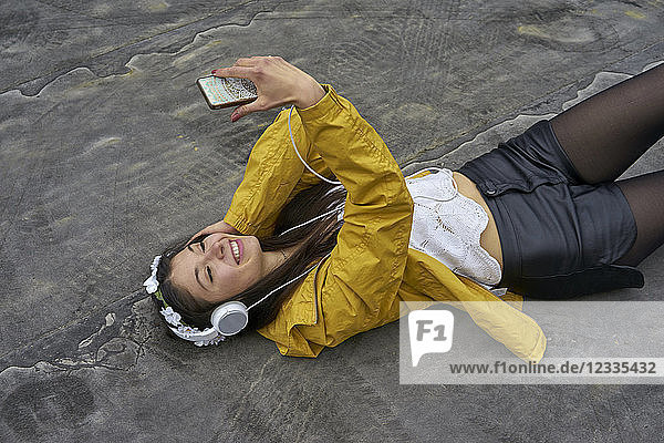 Smiling woman with headphones lying on ground taking selfie with smartphone