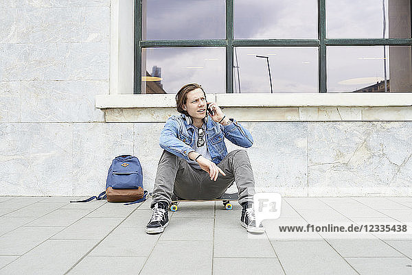 Young man with skateboard sitting on ground  using smartphone