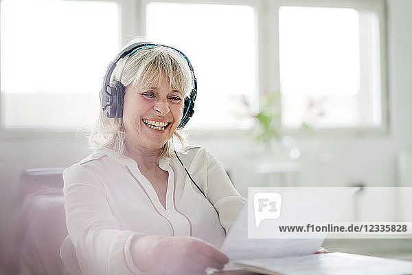Laughing mature businesswoman wearing headphones looking at document at desk