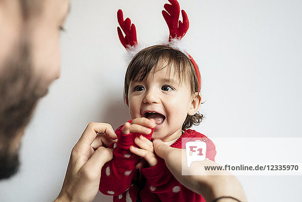 Portrait of laughing baby girl with reindeer antlers headband getting dressed by her father