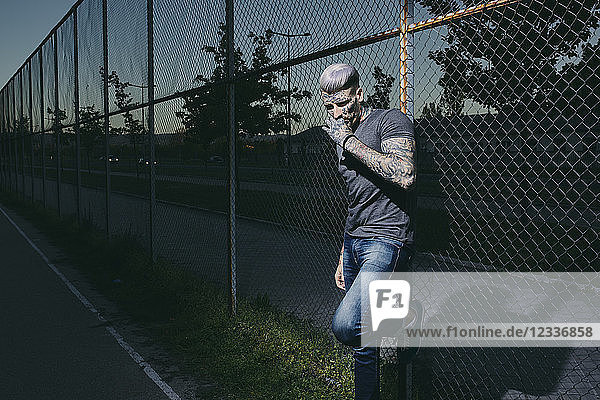 Tattooed young man smoking a cigarette at wire mesh fence