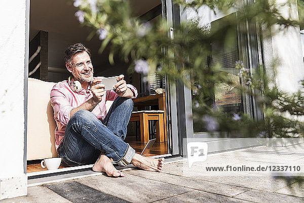 Smiling mature man with smartphone sitting at open terrace door