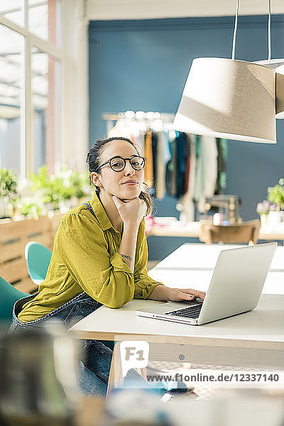 Portrait of fashion designer sitting at desk in her studio with laptop