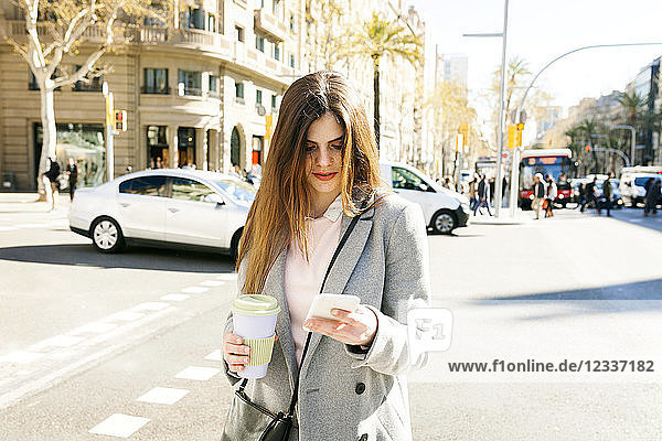 Spain  Barcelona  young woman with coffee to go standing at roadside looking at cell phone