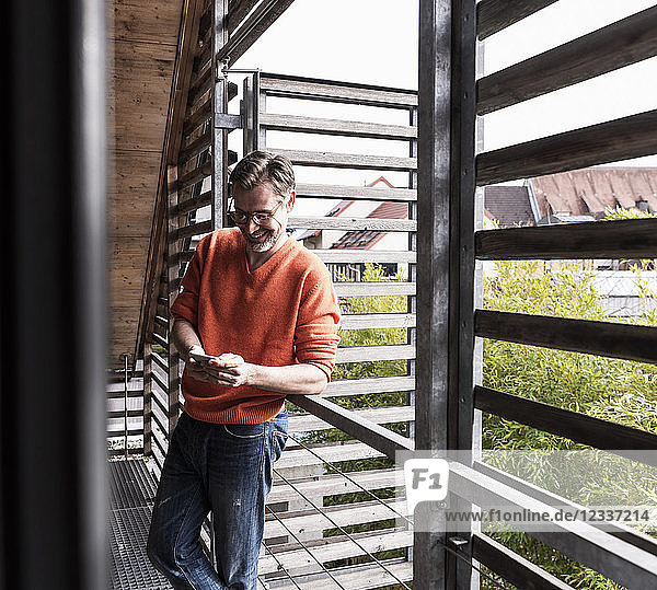 Smiling mature man standing on balcony of his house looking at smartphone
