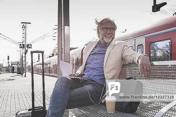 Smiling mature businessman sitting at train station with cell phone  earbuds and notebook