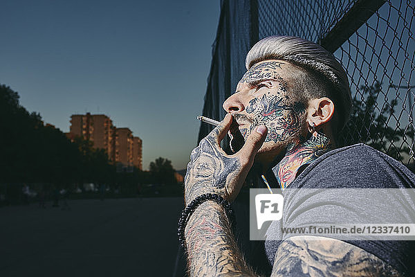Portrait of tattooed young man smoking a cigarette at wire mesh fence