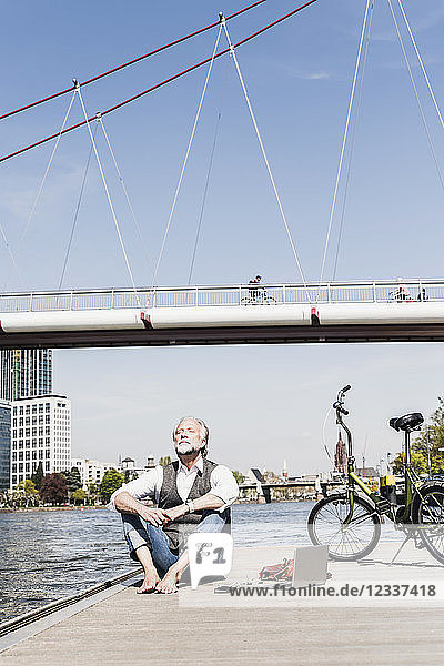 Mature man with laptop and bicycle sitting at the riverside in the city