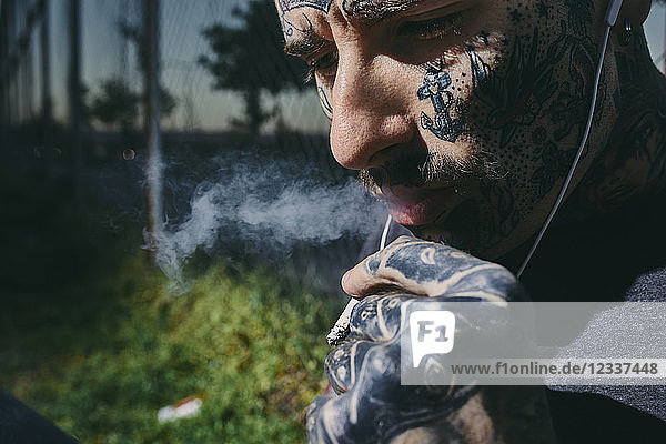 Close-up of tattooed young man with earbuds smoking a cigarette