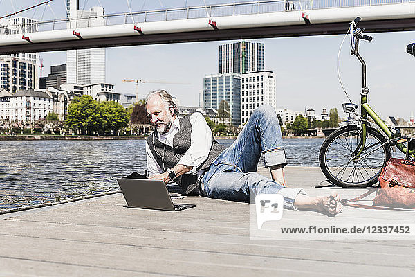 Mature man with laptop  earbuds and bicycle lying at the riverside in the city