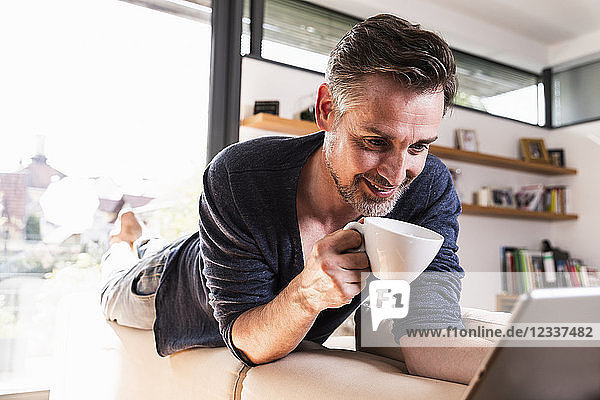 Portrait of smiling man with cup of coffee lying on backrest of couch using tablet