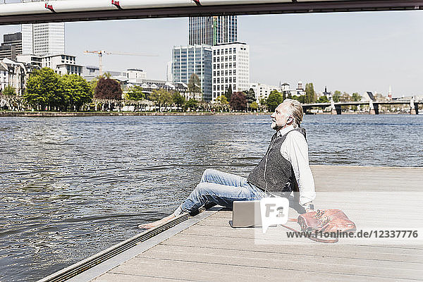 Mature man with laptop and earbuds sitting at the riverside in the city