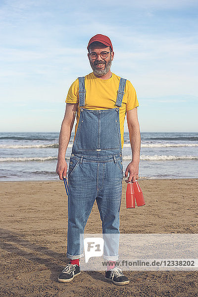 Portrait of smiling man in dungarees standing on the beach holding bottles of soft drinks