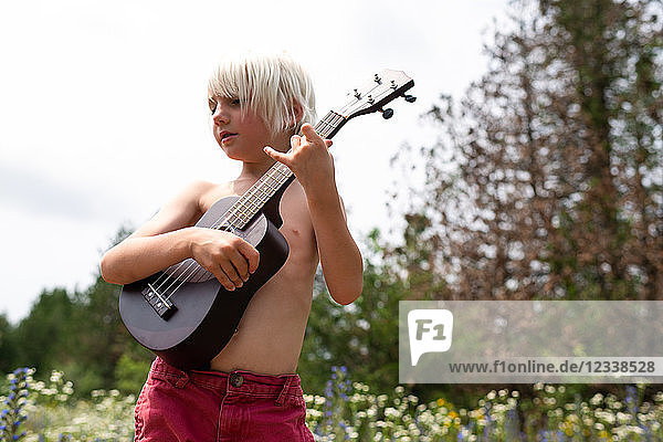 Blondhaariger Junge mit nackter Brust spielt Ukulele auf einer Wildblumenwiese