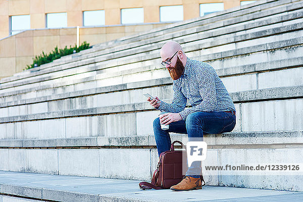 Bärtiger Mann sitzt auf einer Stufe und benutzt ein Smartphone