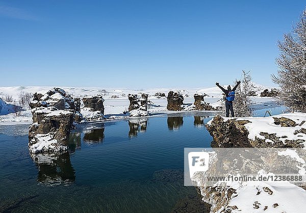 Young man stretches his arms into the air  view of volcanic rock formation in the water  Lake Mývatn in winter  Höfði Peninsula  Iceland  Europe