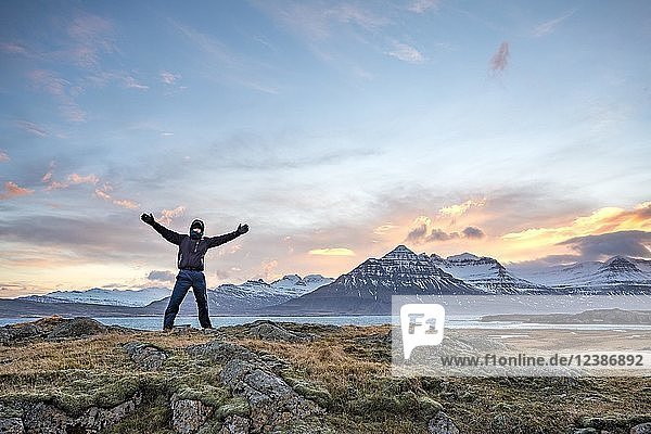 Hiker with outstretched hands standing at the fjord Berufjörður  sunset over snow-covered mountains  Austurland  East Iceland  Iceland  Europe