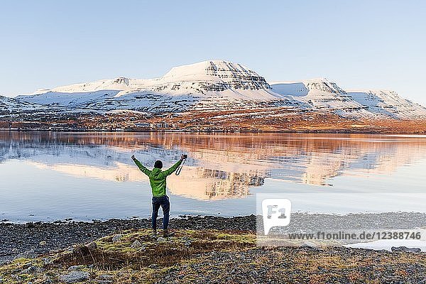 Mann steht am Ufer und streckt die Arme in die Luft  Wasserspiegelung  schneebedeckte Berge in einem Fjord  Reyðarfjörður  Ostisland  Island  Europa