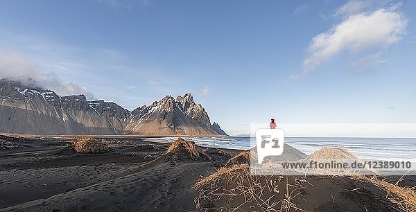 Mann in roter Jacke steht auf Dünen mit trockenem Gras  schwarzer Lavastrand  Sandstrand  Berge Klifatindur  Eystrahorn und Kambhorn  Landzunge Stokksnes  Gebirgszug Klifatindur  Austurland  Ostisland  Island  Europa