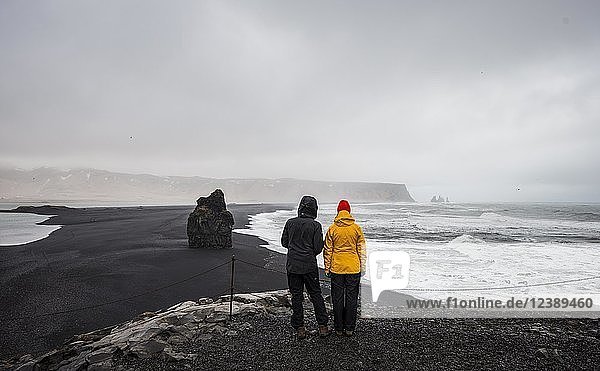 Zwei Touristen mit Blick auf einen schwarzen Sandstrand  schlechtes Wetter  Reynisfjara Strand  Südisland  Island  Europa