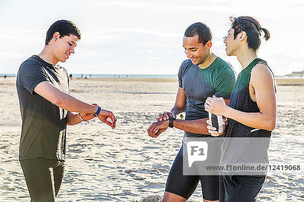 Male runners checking smart watch fitness trackers on sunny beach