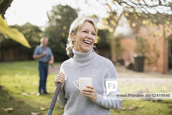 Happy mature woman drinking coffee and raking autumn leaves in backyard