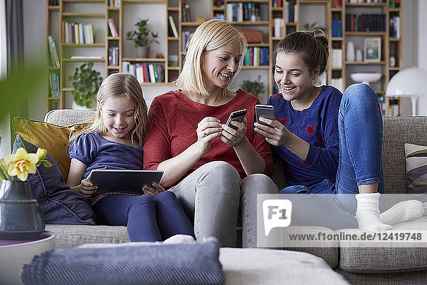 Mother and her daughters sitting on couch  having fun using digital laptop and playing with smartphones
