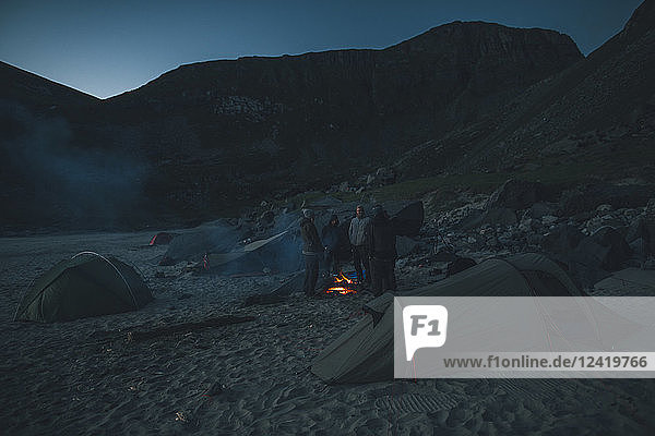 Norway  Lofoten  Moskensoy  Group og young men standing at camp fire on Kvalvika Beach