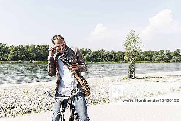 Mature man with bike using smartphone and headphones at Rhine riverbank