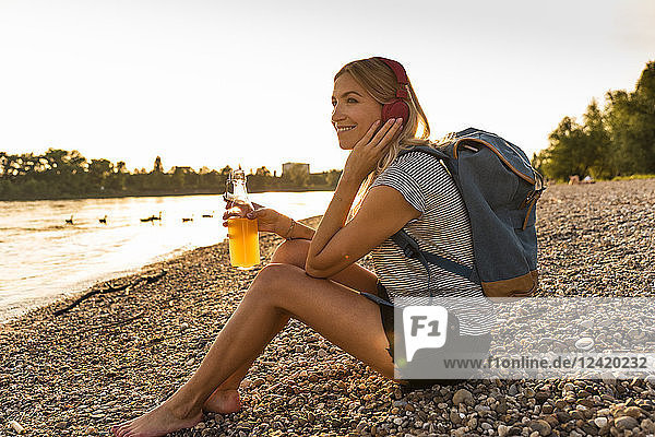 Blond woman with earphone and bottle sitting on riverside in the evening  listening music