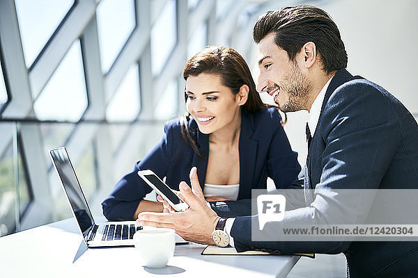 Smiling businesswoman and businessman using laptop and cell phone at desk in modern office