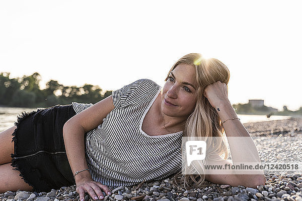 Young blond woman lying at riverside against evening sun