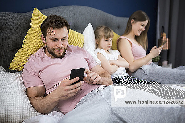 Neglected little girl sitting on bed with her parents  using smartphones