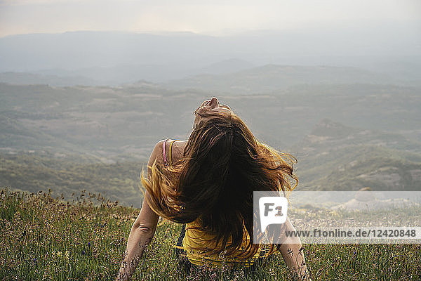 Spain  Barcelona  back view of young woman sitting on meadow tossing her hair