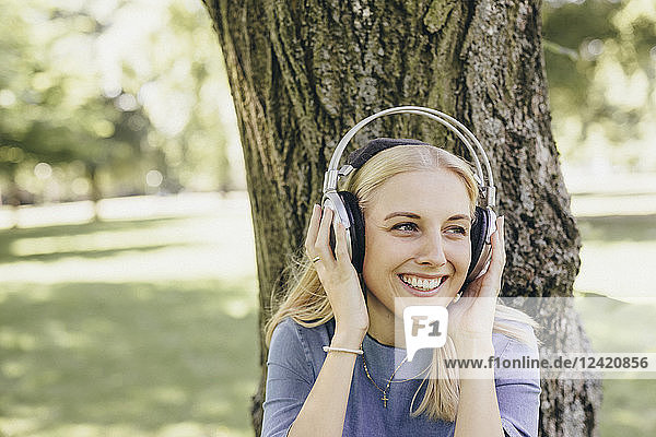 Happy young woman at tree trunk in a park wearing headphones