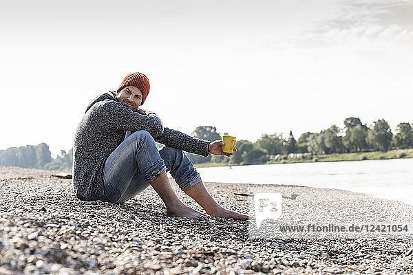 Mature man wearing red beanie and drinking coffee at Rhine riverbank