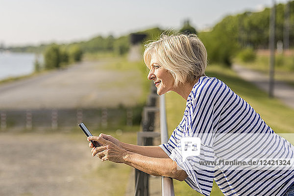 Smiling senior woman with cell phone leaning on railing at riverbank