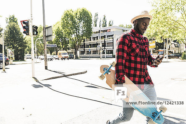 Cool young man with skateboard walking in the city  using smartphone
