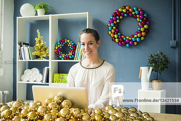 Portrait of smiling woman sitting at table with laptop and many golden Christmas baubles